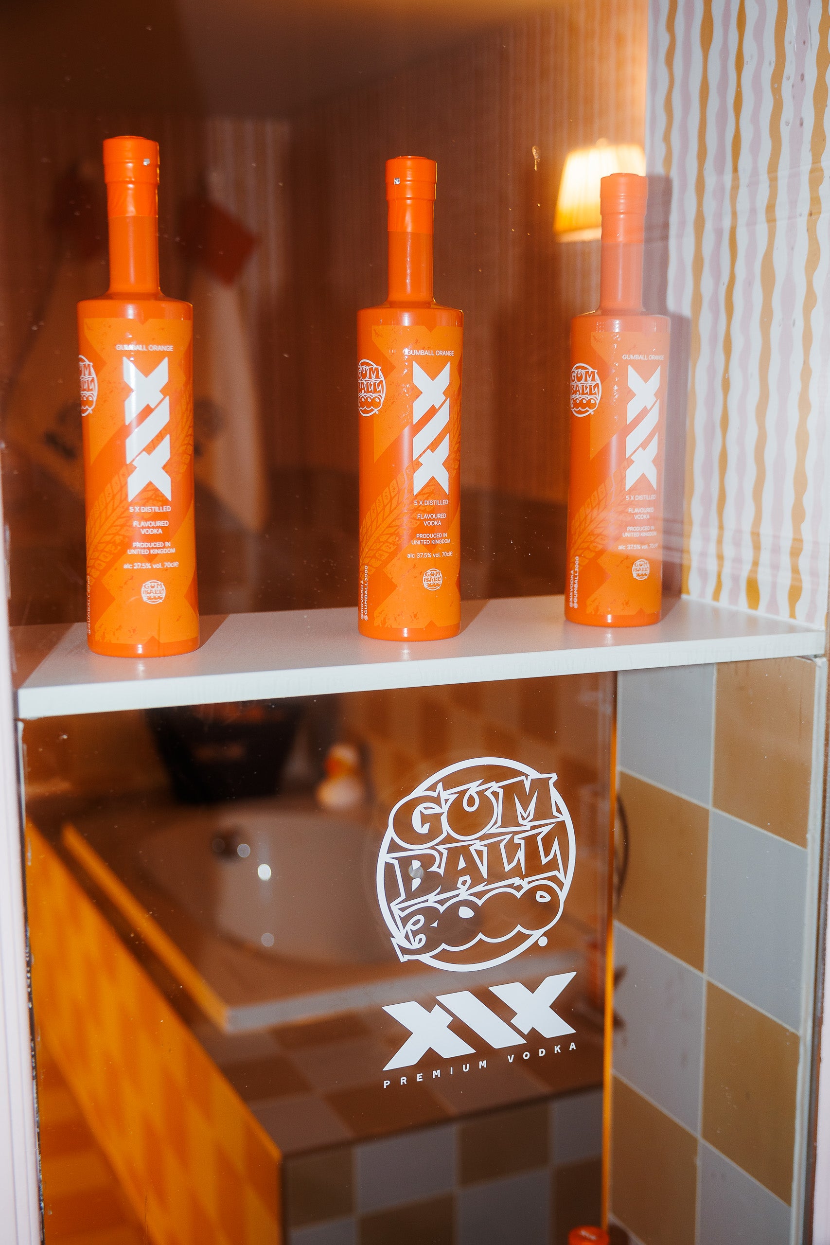 Three orange bottles on a shelf with a branded glass panel in the background.