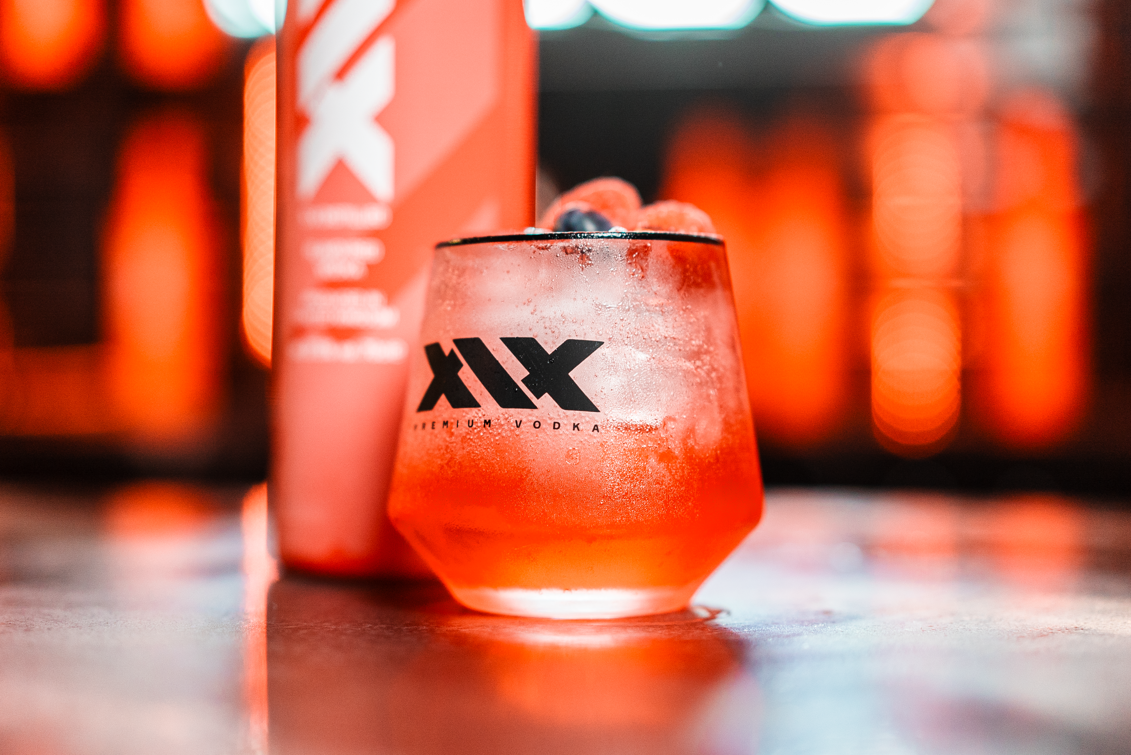 Glass of red cocktail with ice and a bottle of XIV Vodka on a reflective surface with blurred lights in the background.
