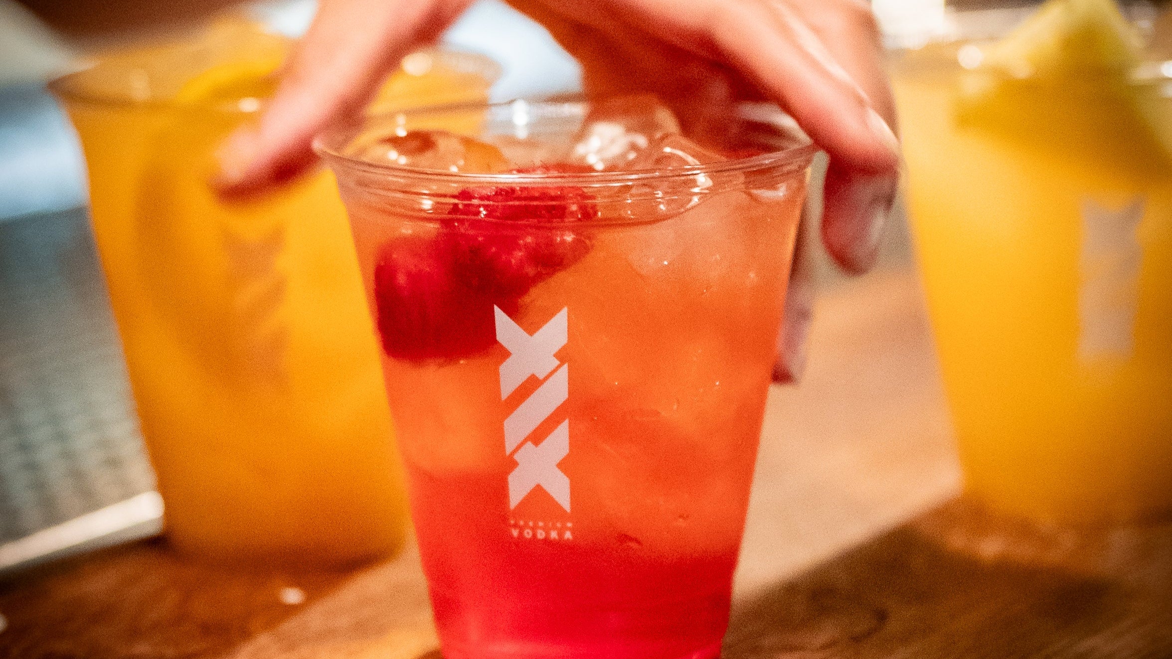 Three drinks in clear plastic cups with a logo on a wooden surface, one of which is being picked up.