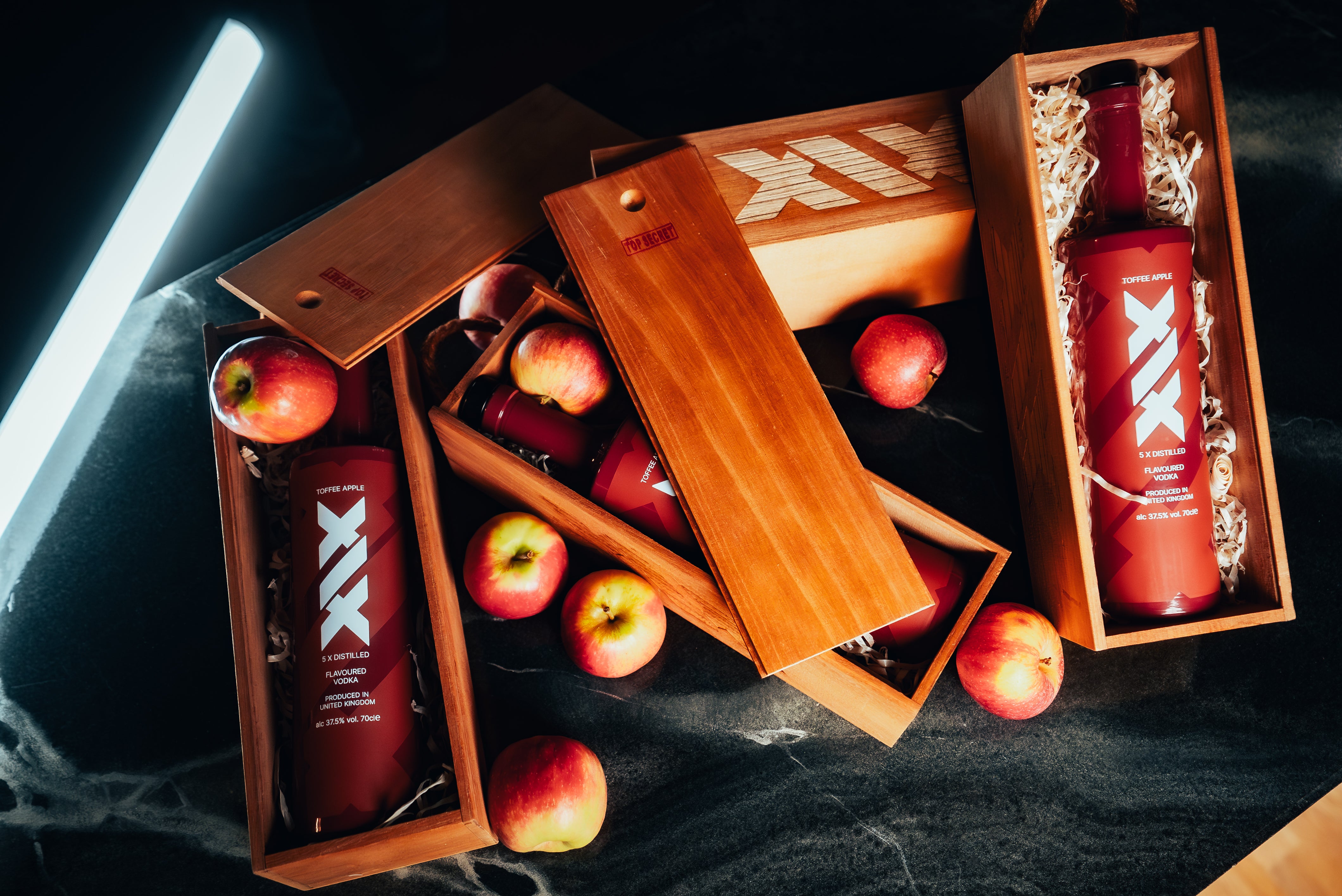 Brown bottles with 'XX' branding in wooden gift boxes surrounded by apples on a dark surface.